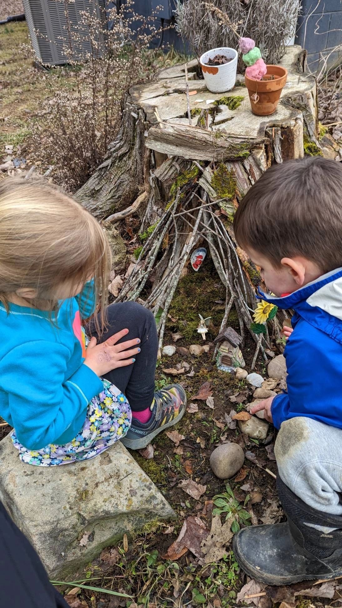Two children explore a miniature fairy house built at a tree stump.